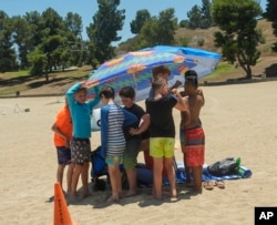 Youths shelter in the shade after being asked by lifeguards to retrieve their footwear before getting onto the sunny beach area to prevent burned feet at Castaic Lake beach as the temperature rises, July 8, 2024, in Castaic, Calif.