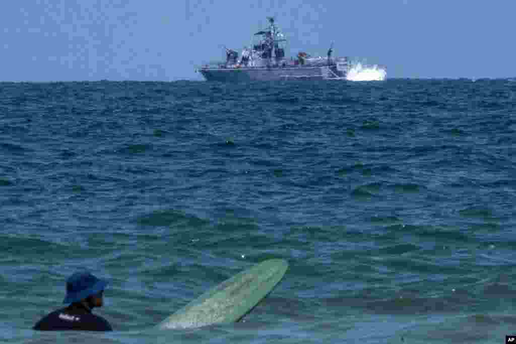 A surfer waits for wave while an Israeli military naval ship patrols the Mediterranean sea off the coast of Hadera, Israel, April 14, 2024.