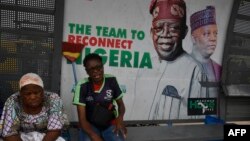 FILE - Passengers sit at a bus station beside a billboard of President Bola Tinubu and Vice President Kashim Shettima, while waiting at a bus station in Lagos, June 1, 2023. Nigerian consumers are lamenting the ripple effect of the removal of the fuel subsidy.