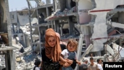 Seorang perempuan Palestina menggendong putrinya saat berjalan melewati puing-puing rumah yang hancur akibat serangan militer Israel, di Khan Younis di selatan Jalur Gaza, 10 Juli 2024. (REUTERS/Hatem Khaled)