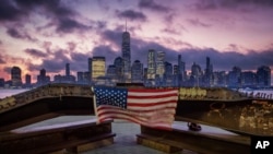 FILE - A U.S. flag hanging from a steel girder, damaged in the Sept. 11, 2001 attacks on the World Trade Center, blows in the breeze at a memorial in Jersey City, N.J., Sept. 11, 2019 as the sun rises behind One World Trade Center building in New York.
