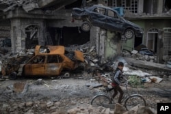 FILE - A boy rides his bike past destroyed cars and houses in a neighborhood recently liberated by Iraqi security forces on the western side of Mosul, Iraq, March 19, 2017.