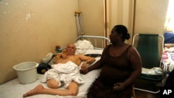 In this Aug. 24, 2015, photo, Bertha More, 54, sits with her husband Alberto Espinosa, 79, a former restaurant worker who was paralyzed by a stroke seven years ago and later lost a leg to circulation problems, in Havana, Cuba.