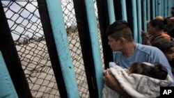 A member of the Central American migrant caravan, holding a child, looks through the border wall toward a group of people gathered on the U.S. side, as he stands on the beach where the border wall ends in the ocean, in Tijuana, Mexico, April 29, 2018. 