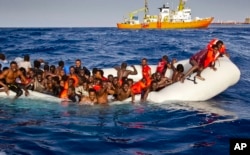 FILE - Migrants ask for help from a dinghy boat as they are approached by the SOS Meditrranee's ship Aquarius, background, off the coast of the Italian island of Lampedusa, April 17, 2016.