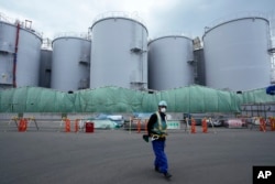 Seorang pekerja membantu mengarahkan pengemudi truk saat dia berdiri di dekat tangki yang digunakan untuk menyimpan air radioaktif yang diolah setelah digunakan untuk mendinginkan bahan bakar di PLTN Fukushima Daiichi, di kota Okuma, timur laut Jepang, Kamis, 3 Maret, 2022. (Foto: AP)
