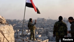Under the Syrian flag, a government soldier makes a V sign while overlooking eastern Aleppo, after troops took control of the city's al-Sakhour neigborhood, in this photo provided by SANA, the state-run news agency, Nov. 28, 2016.