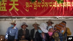 পশ্চিম চীনের তিব্বত স্বায়ত্তশাসিত অঞ্চলের লাসার চেংগুয়ান জেলার একটি পার্শ্ববর্তী তিব্বতি বৌদ্ধ মন্দিরের কাছে চীনা এবং তিব্বতি ভাষায় একটি সরকারি প্রচার ব্যানারের নীচে কয়েকজনকে বসে থাকতে দেখা যাচ্ছে। ৩ জুন, ২০২১। ফাইল ছবি। 