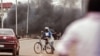Smoke rises as Burkina Faso troops, in rear, clear debris from a road as people protest in the area against the recent coup in the city of Ouagadougou, Burkina Faso, Sept. 18, 2015.
