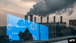 FILE - Pedestrians and an electronic screen displaying COVID-19 prevention advice is reflected in a glass wall as smog billows from a factory in Moscow, Dec. 3, 2020.