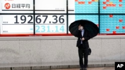 A man wearing a protective mask stands in the rain in front of an electronic stock board showing Japan's Nikkei 225 index at a securities firm in Tokyo Monday, April 13, 2020.