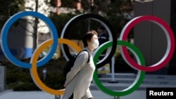FILE - A woman wearing a protective face mask, following an outbreak of the coronavirus disease (COVID-19), walks past the Olympic rings in front of the Japan Olympics Museum in Tokyo, March 13, 2020.