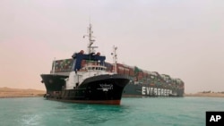 A boat navigates in front of a massive cargo ship, named the Ever Green, rear, sits grounded March 24, 2021, after it turned sideways in Egypt’s Suez Canal, blocking traffic in a crucial East-West waterway for global shipping.