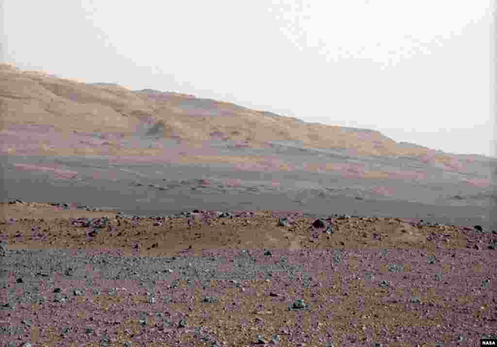 The gravelly area around Curiosity's landing site is in the foreground. Beyond the swale is the red-brown rim of an impact crater. Further in the distance, there are dark dunes and the layered rock at the base of Mount Sharp.