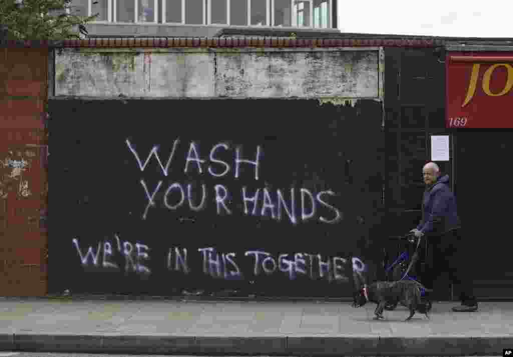 Un hombre pasa a su perro cerca de una pared con un graffiti que pide a las personas que se laven las manos, en Belfast, Irlanda del Norte.