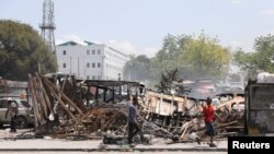 হেইতির রাজধানী পোর্ট অ প্রিন্স এ গ্যাং সহিংসতায় বিধ্বস্ত যানবাহনের পাশ দিয়ে লোকজন হেঁটে যাচ্ছেন। ফটোঃ ২৫ মার্চ, ২০২৪। 