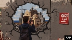 အစ္စရေးတို့ ကာရံထားတဲ့ နယ်စပ်တံတိုင်းရိုးကြီးပေါ်မှာ နံရံဆေးမှုတ်အနုပညာနဲ့ ပုံဖေါ်ထားတဲ့ ဂါဇာ ပြန်လည်ထူထောင်တည်ဆောက်ရေး စိတ်ကူးပုံရိပ်ကို လမ်းသွားတဦး သူ့ဖုန်းနဲ့ ဓါတ်ပုံရိုက်ယူနေတာလေ။ (February 12, 2025)