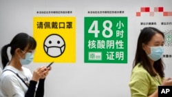FILE - People wearing face masks stand near signs reminding them to wear a mask and show proof of a negative COVID test within the past 48 hours as they wait to enter an exhibition at a shopping mall in Beijing, Sept. 24, 2022.