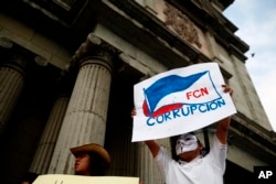 FILE - A masked man holds up a sign that reads "FCN Corruption," referring to the ruling party, Frente Convergencia Nacional, as he and others protest Guatemala's President Jimmy Morales at Constitution Square in Guatemala City, April 21, 2018.
