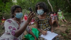 FILE - A Sri Lankan woman holds her smart phone for her daughter to take online classes from a signal reception point on a mountain in a reserve forest in Lunugala, Sri Lanka, July 3, 2021.