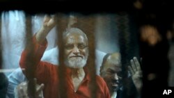 Pemimpin Ikhwanul Muslimin, Mohammed Badie mengenakan seragam merah yang mengindikasikan bahwa ia dijatuhi hukuman mati, melambai dari bilik terdakwa di pengadilan darurat di akademi polisi nasional, Kairo timur, Mesir, Sabtu, 16 Mei 2015. (AP Photo/Ahmed Omar)