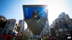 FILE - In this April 26, 2019, photo, traffic warden and securities stand guard near a TV screen broadcasting live of President Xi Jinping's opening speech, outside a shopping mall in Beijing.
