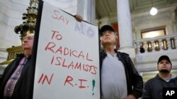Howard Brown, of North Kingstown, R.I., center, displays a placard during a rally at the statehouse in Providence, R.I., held to demonstrate against allowing Syrian refugees to enter Rhode Island, Nov. 19, 2015. 