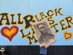FILE - A demonstrator carries an image of George Floyd in front of a boarded-up business decorated with a mural reading "All Black Lives Matter," during a march on Hollywood Boulevard in Los Angeles, June 14, 2020.