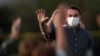 Presiden Brazil Jair Bolsonaro yang terinfeksi Covid-19, mengenakan masker saat melambaikan tangannya kepada para suporternya yang menghadiri upacara penurunan bendera di luar kediaman resminya Istana Alvorada, di Brasilia, Brasil, Senin, 20 Juli 2020. (Foto AP / Eraldo) 