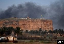 This picture taken on March 22, 2019 shows smoke rising over the village of Baghuz in the eastern Syrian province of Deir Ezzor, with the flags of the Kurdish People's Protection Units (Yellow) and Women's Protection Units (Green) seen flying.