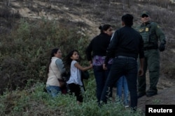 Migrants, part of a caravan of thousands from Central America trying to reach the United States, stand next to a U.S. Customs and Border Protection official in San Diego County, after crossing illegally from Mexico, Dec. 2, 2018.