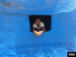 A displaced Sunni teenager looks out of a window cut into plastic blue sheeting covering his new home in the Baharka camp on the outskirts of Irbil, in the Kurdistan region of Iraq, Aug 5, 2015. (VOA / S.Behn)