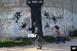 FILE - Palestinian children walk next to a mural of children using an Israeli army watch tower as a carnival ride, presumably painted by British street graffiti artist Banksy, on the main road in Beit Lahiya, in the northern Gaza Strip, Friday, Feb. 27, 2