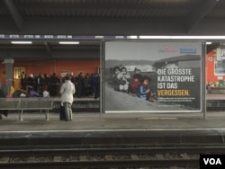 Migrants await their fate at Freilassing's train station in Germany near a billboard by charity groups urging Germans to support refugees. It reads: "The biggest catastrophe is forgetting. 12 million people from Syria and Iraq are fleeing. We must support them." (L. Ramirez/VOA)