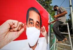 FILE - A worker sits on a ladder as he installs a coronavirus awareness-themed banner bearing a portrait of Indonesian President Joko Widodo wearing a face mask, in Jakarta, Indonesia, Aug. 26, 2020.