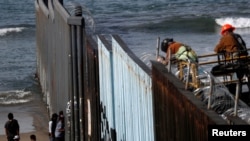 Migrants, part of a caravan of thousands trying to reach the U.S., stand by the border fence between Mexico and the United States, as a man welds concertina wire, as seen from Tijuana, Mexico Nov. 15, 2018. 