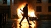 A man with a stick in hand walks past a burning police car not far from the Georgian parliament building in Tbilisi, Georgia, March 9, 2023. 