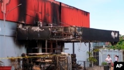 Seorang perempuan membawa barang belanjaannya berjalan di dekat klub malam yang terbakar di Sorong, provinsi Papua Barat, Selasa, 25 Januari 2022. Api melalap klub malam setelah dua kelompok bentrok di dalam gedung tersebut. (Foto AP)