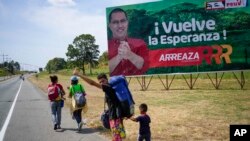 Una familia de venezolanos que emigra a Colombia hace autostop frente a una valla publicitaria que invita a votar por el candidato a gobernador del partido gobernante, Jorge Arreaza, en Barinas, Venezuela, el viernes 21 de enero de 2019. 