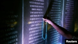 FILE - Margaret Achieng Jow, whose daughter was killed in the US. Embassy in Nairobi bombing, holds a cross on a wall displaying the names of people killed during the attack, at a ceremony marking the 20th anniversary of the bombing, Aug. 7, 2018.