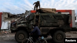 A man wearing a protective face mask rides a bike past a soldier on an American MaxxPro military vehicle in the formerly Russian occupied city of Lyman, Donetsk region of Ukraine, Dec. 11, 2022. 