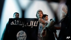 Dalam foto dokumentasi dari tahun 2013 ini, seorang yang diduga sebagai militan al-Qaida di Yaman, tengah, membentangkan bendera kelompok ISIS di tengah berlangsungnya sebuah persidangan di Sana'a, Yaman. Dalam persaingannya dengan kelompok Negara Islam (ISIS) di Timur Tengah, al-Qaida menggunakan pendekatan yang dianggap sebagian kalangan sebagai pragmatis.