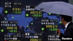 A pedestrian holding an umbrella looks at an electronic board the stock market indices of various countries outside a brokerage in Tokyo, Japan, July 6, 2015. 