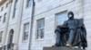 FILE - The John Harvard statue at Harvard University, a popular tourist attraction at the campus in Cambridge, Masssachusetts, sits adorned with a mask, March 14, 2020. The university is restricting spending in the wake of the coronavirus outbreak.