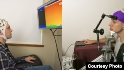 UW students Darby Losey, left, and Jose Ceballos are positioned in two different buildings on campus as they would be during a brain-to-brain interface demonstration. The sender, left, thinks about firing a cannon at various points throughout a computer game. That signal is sent over the Web directly to the brain of the receiver, right, whose hand hits a touchpad to fire the cannon.Mary Levin, U of Wash.